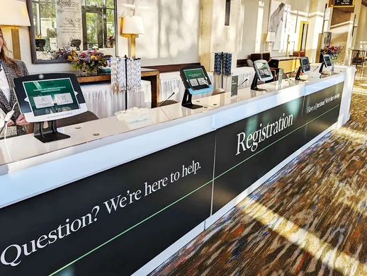 A staffed registration desk at a life sciences event: self check-in tablets and badge printers on the counter, ready for arrival.